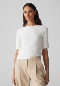 Witte geribbelde top met korte mouwen, ronde halslijn en een getailleerde pasvorm, gecombineerd met beige hoge taille broeken. Soepele textuur, minimalistisch ontwerp.