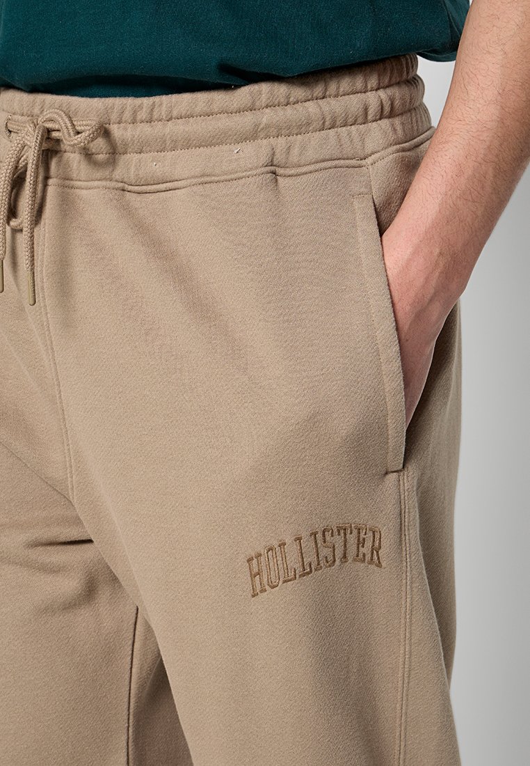 Beżowe dresy wykonane z miękkiego materiału, z elastycznym pasem na sznurki, bocznymi kieszeniami oraz wytłoczonym logo "HOLLISTER" na nodze.