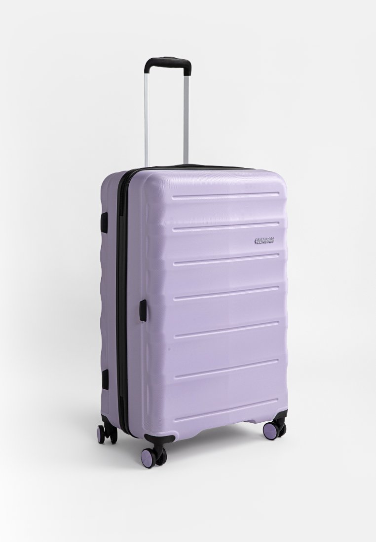 Valise rigide de couleur lavande, avec une texture striée, fermeture éclair noire et poignée télescopique. Quatre roues pour la mobilité.