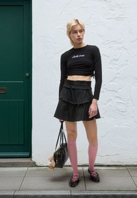 Crna kratka majica s dugim rukavima i ljubičastim tekstom, slojevita crna naborana suknja, ružičaste čarape do koljena i crna torbica s privjeskom.