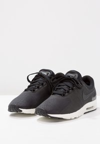 Mustat urheilukengät, joissa on tekstuuri päällä, nauhakiinnitys ja valkoinen pohja. Ominaisuuksiin kuuluvat näkyvä ilmayksikkö ja Nike-logo sivussa.