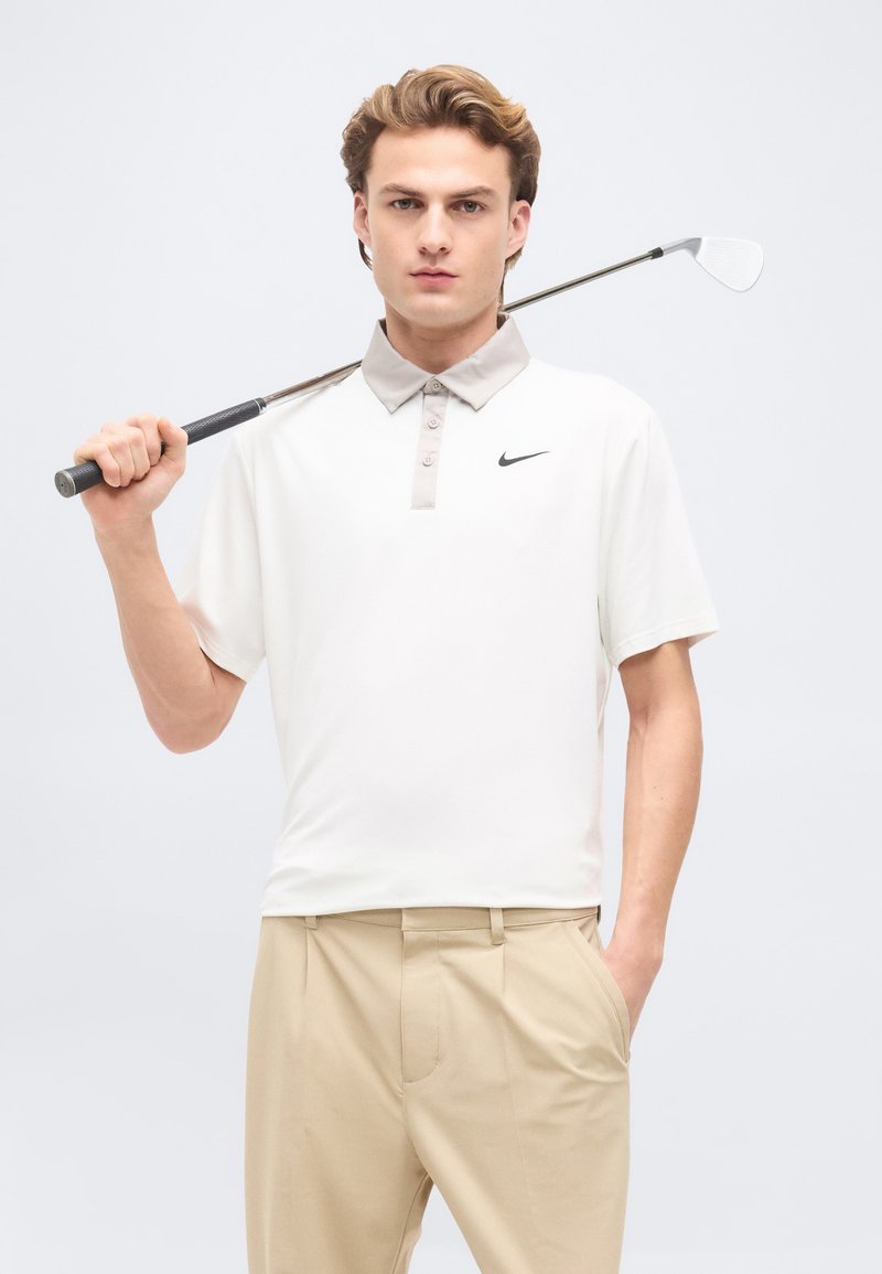 Jeune homme en pantalon beige et polo blanc tenant un club de golf derrière son cou sur un fond uni gris clair.