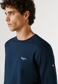 Ung mand med krøllet hår iført en mørkeblå langærmet Pepe Jeans London skjorte, set fra siden mod en ensfarvet lys baggrund.