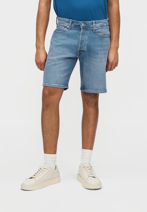 Man die lichtblauwe denim shorts, witte sokken en witte sneakers draagt, staande tegen een effen achtergrond.