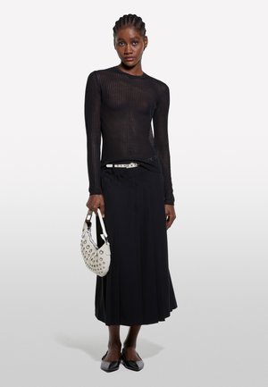 Donna con capelli intrecciati che indossa una maglia nera trasparente a maniche lunghe, gonna nera, scarpe nere, cintura bianca e tiene in mano una borsa bianca con borchie.