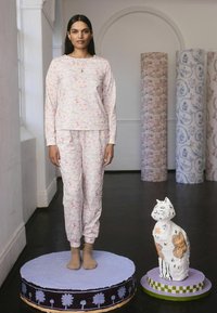 Een vrouw in een patroon pyjama staat op een rond platform naast een geschilderde kat sculptuur, omringd door decoratieve kolommen.