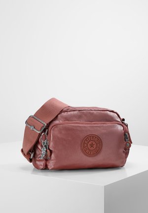 Petit sac bandoulière rose poussiéreux avec une sangle réglable, une poche zippée à l'avant et un patch rond avec le logo Kipling, posé sur un piédestal blanc.