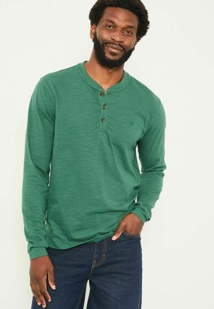Grünes Langarm-Henley-Shirt aus strukturiertem Stoff mit drei Knöpfen am Hals, kombiniert mit dunkelblauen Jeans. Einfaches, lässiges Design.