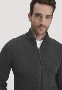 Grauer Reißverschluss-Pullover mit geripptem Kragen, Strickstruktur undmetallischem Reißverschluss. Verfügt über ein tailliertes Design und wird über einem weißen Shirt getragen.