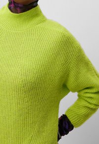 Pull tricoté vert citron avec un col montant, une texture côtelée et une coupe ample. Les détails comprennent des manches larges et une silhouette légèrement oversize.