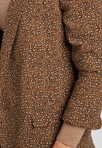 Giacca marrone con stampa leopardata, realizzata in un tessuto testurizzato, dotata di due tasche frontali e dettagli a costine sui polsini. Il motivo include accenti neri e bianchi.