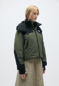 Jaqueta verde e preta com gola alta, bolsos com fecho éclair e tecido texturizado. As punhos têm detalhes em preto; o design é moderno e funcional.