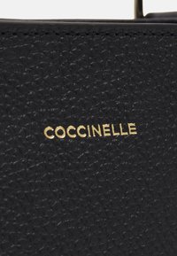 Sac en cuir noir texturé avec un logo "Coccinelle" doré. La surface présente un motif distinct, ajoutant un intérêt visuel au design.