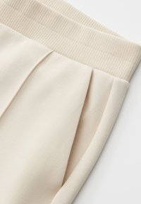 Beige pants with ribbed elastic waistband, front pleat, and side pocket, made of smooth fabric.