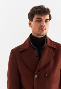 Brązowy wełniany płaszcz dwurzędowy z dużymi klapami, zestawiony z czarnym swetrem z wysokim kołnierzem. Gładka tekstura i dwa czarne guziki na płaszczu.