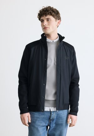 Fiatal férfi rövid, göndör hajjal, fekete cipzáras dzsekit visel világosszürke pulóver felett és kék farmert, enyhén jobbra néz.