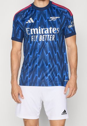 Mann trägt blaues Arsenal-Fußballtrikot mit Zickzackmuster, weißem Emirates-Sponsortext und weißen Adidas-Shorts.