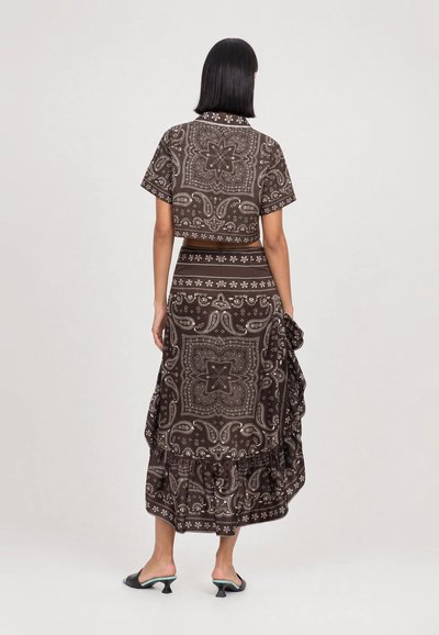 Femme aux cheveux noirs et raides porte un haut marron à manches courtes avec un motif paisley et une longue jupe assortie avec un détail volanté, vue de dos.