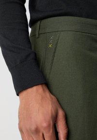 Pantalons ajustés verts avec un devant plat et une couture jaune d'accent. Associé à un pull noir côtelé. Vue rapprochée mettant en valeur la texture du tissu.