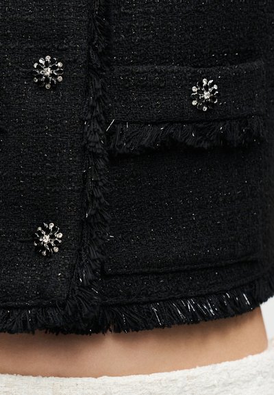 Μαύρο υφασμάτινο με υφή, με κρόσσια στις άκρες και λαμπερά ασημί κουμπιά σε σχήμα λουλουδιού σε τσέπη ενδύματος.