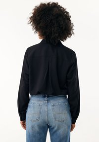 Černá halenka s dlouhým rukávem a límečkem, vybavená středním záševem na zadní straně. Spárována s modrými džíny s vysokým pasem.