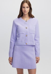Lila Strickjacke aus strukturiertem Stoff, mit zwei vorderen Taschen und goldfarbenen Knöpfen. Kombiniert mit einem passenden lila Rock.