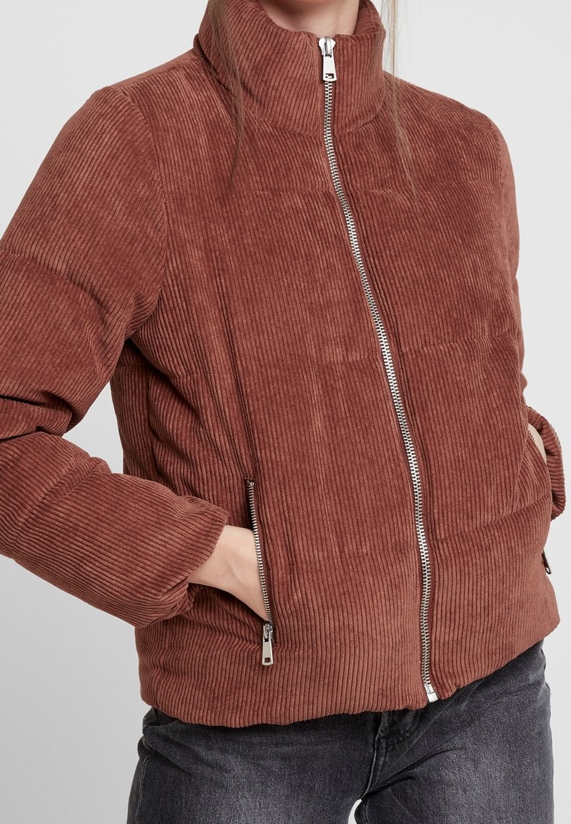 Hnědá kordurová pufová bunda s vysokým límcem, předním zipem a bočními kapsami. Vyznačuje se vertikálním ribbovaným vzorem a elastickými manžetami.