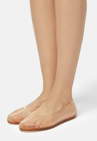 Feet wearing translucent, peach-colored woven ballet flats with a rounded toe, shown against a plain white background.