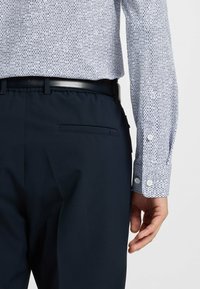 Vit skjorta med blå mönster, långa ärmar och knappmanschetter; mörk marinblå byxor med en slät yta och bakfickdetalj.