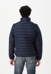 Jachetă puf cu design matlasat orizontal, de culoare navy, cu guler înalt și manșete elastice, purtată cu blugi de culoare bleu deschis.