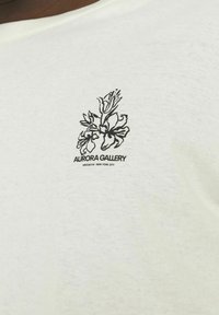 T-shirt en coton blanc avec une illustration florale noire et le texte "AURORA GALLERY BROOKLYN, NEW YORK CITY" en dessous du design.
