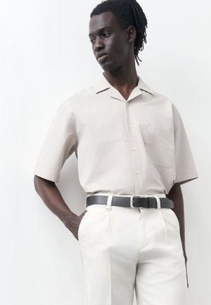 Mężczyzna o ciemnej skórze i dredach, ubrany w jasnobeżową koszulę z krótkim rękawem zapinaną na guziki oraz białe spodnie z czarnym paskiem, pozujący na białym tle.