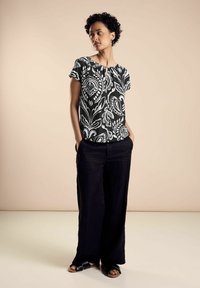 Top a maniche corte a scacchi bianco e nero con scollo a giro, abbinato a pantaloni neri larghi e sandali neri piatti, su sfondo beige.