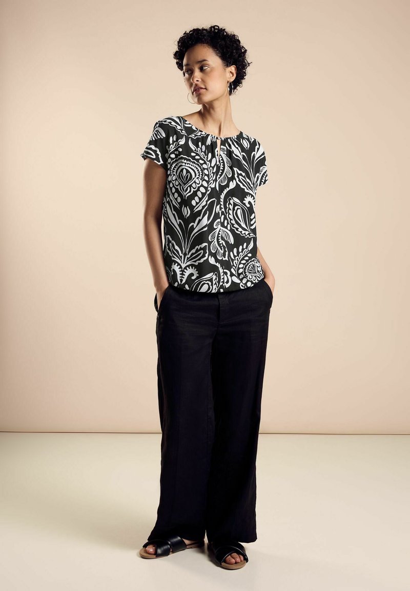 Top a maniche corte a scacchi bianco e nero con scollo a giro, abbinato a pantaloni neri larghi e sandali neri piatti, su sfondo beige.