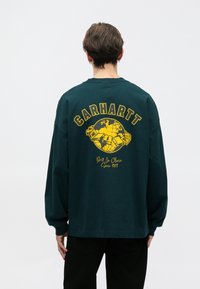 Green long-sleeve shirt featuring a large yellow graphic on the back with text and a globe design. Cotton material with a relaxed fit.