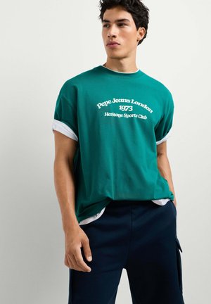 Junger Mann trägt ein türkisfarbenes Pepe Jeans London T-Shirt mit weißem Schriftzug und marineblaue Hose, steht mit einer Hand in der Tasche vor einem schlichten Hintergrund.
