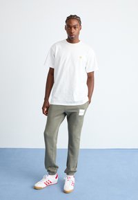 Tricou alb din bumbac cu un mic logo galben pe piept, combinat cu pantaloni de trening gri care au un patch cu logo alb, adidași albi cu accente roșii.