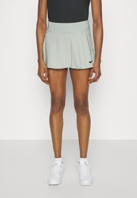 Falda deportiva verde claro con una cinturilla ancha y diseño plisado. Combinada con zapatillas blancas y una camiseta negra ajustada. Logo de Nike en la falda.