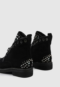 Botines de ante negro con acabado texturizado, que presentan tachuelas plateadas y acentos decorativos en los tacones. Cierre con cremallera lateral.