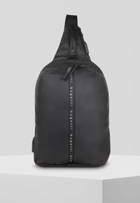 Black sling bag with a smooth, matte finish and textured strap. Features a central zipper and branding down the side in white letters.