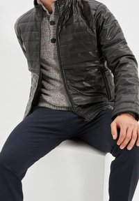 Uomo seduto su un blocco bianco che indossa una giacca trapuntata nera, un maglione grigio a maglia con bottoni e pantaloni scuri.