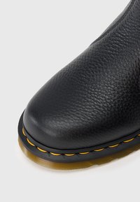 Bota de couro preta com acabamento texturizado, bico arredondado e costura amarela numa sola de borracha. O design apresenta um estilo slip-on.