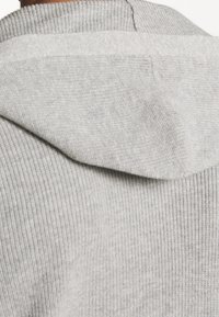 Šedá pletená mikina s kapucí, která má vroubkovanou texturu, podšitý límec a kontrastní vertikální prošívání. Hladký materiál s jemným povrchem.