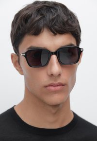 Occhiali da sole rettangolari neri con lenti scure, caratterizzati da un design elegante e accenti metallici sulle cerniere. Indossati da un modello con capelli corti.