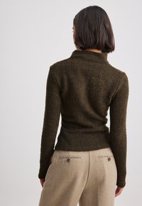 Ruskea tekstuuri neule, jossa on korkea kaulus, tiukka malli ja pitkät hihat. Yhdistetty beigeihin housuihin, joissa on takataskut ja nappikoristeet.