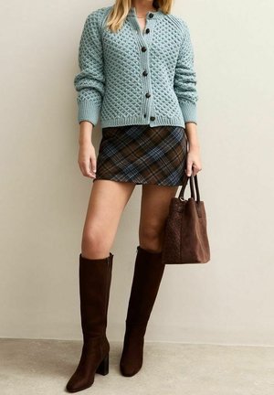 Femme portant un cardigan en tricot bleu boutonné, une mini-jupe marron à carreaux, des bottes marron foncé montantes et tenant un sac à main en daim marron.
