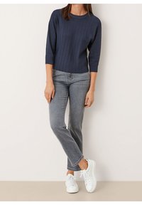 Femme portant un pull côtelé bleu marine à manches trois-quarts, un jean droit gris et des baskets blanches, debout devant un fond clair uni.