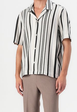 Homme portant une chemise à manches courtes, boutonnée, à rayures verticales noires et blanches, associée à un pantalon marron clair, debout.