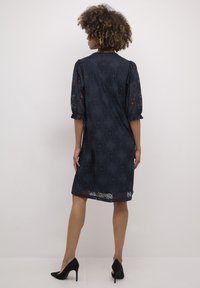 Tengerészkék csipkeblúz kis puffos ujjakkal, virágmintákkal, laza fazonnal és térd alatti szegéllyel. Fekete magassarkú cipők.