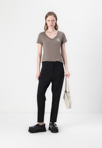 Rudas V formas dekoltė marškinėliai su baltu "CK" logotipu, derinami su juodomis pritaikytomis kelnėmis ir masyviais juodais batais. Modelis laiko šviesiai beige spalvos rankinę.